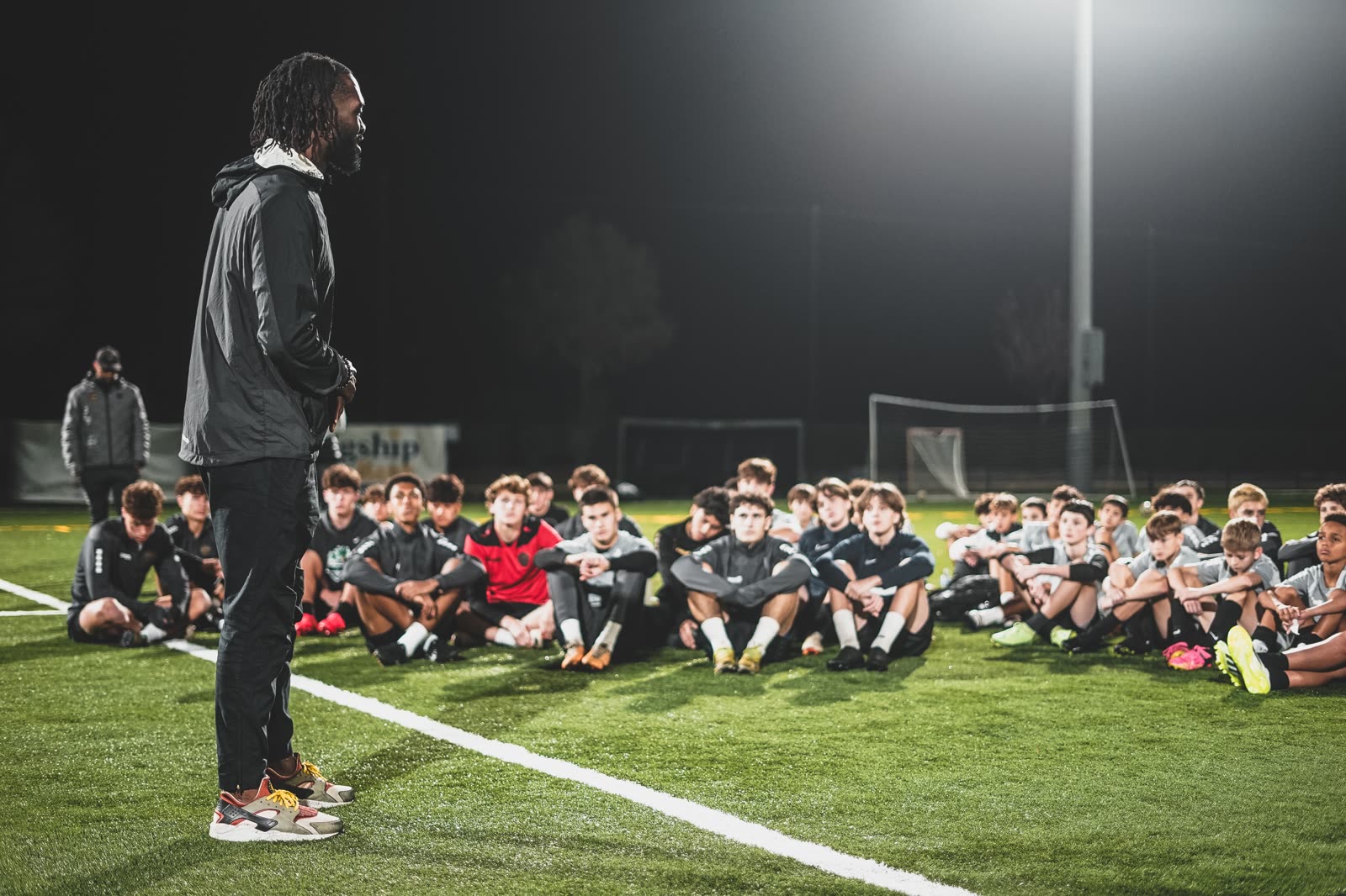 CJ Sapong coaching, standing in front of a large group of young players seated on a floodlit pitch.